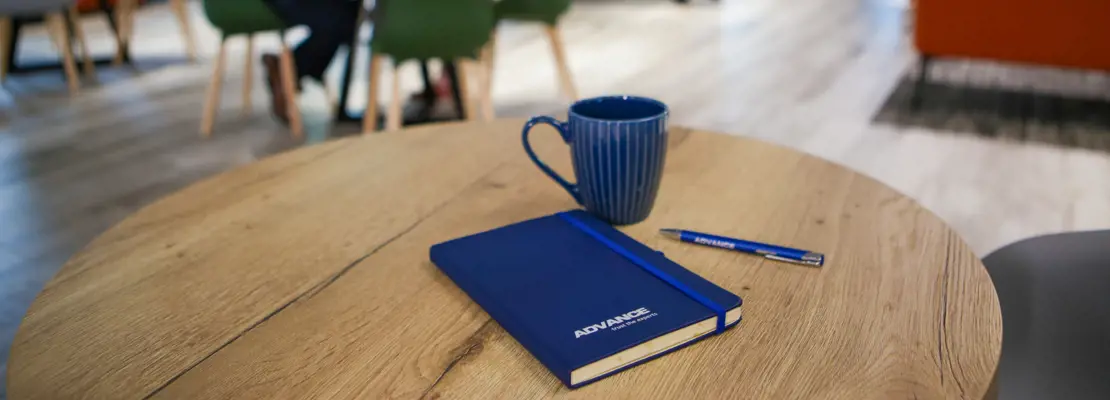 A photo of an ADVANCE Mug with a ADVANCE Pen and Notebook on a table