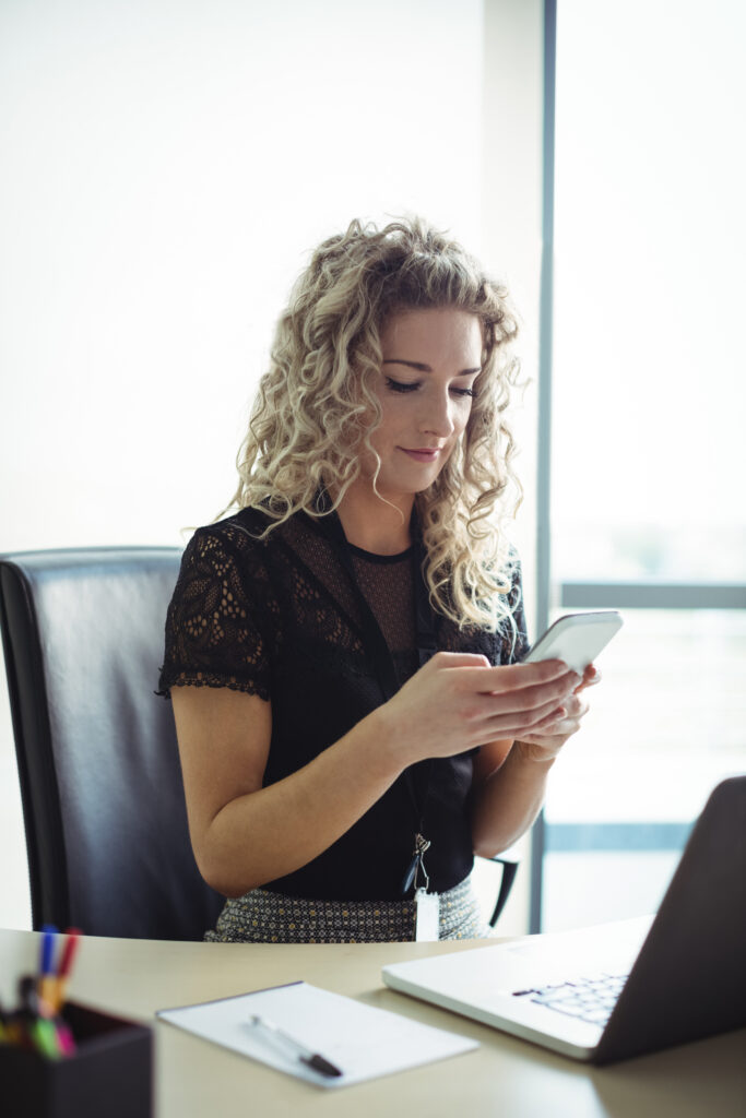 female executive smiling looking at her phone in her office with her laptop open