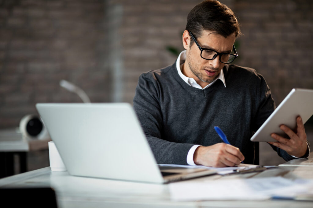 Man smartly dressed coping notes from a notepad whilst sat at his desk with his laptop open,