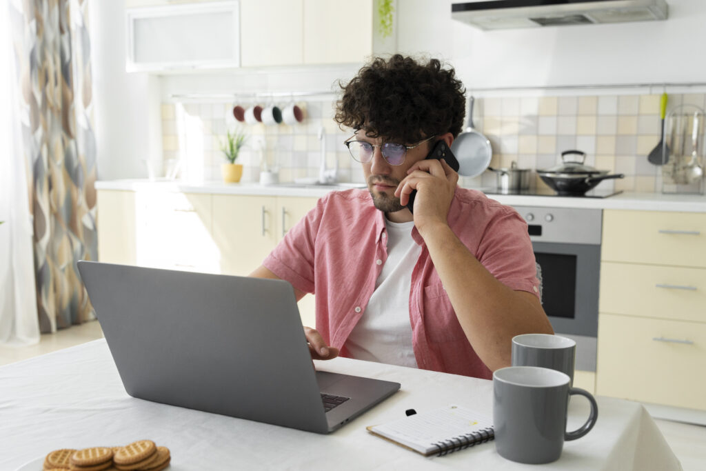 Man sitting in the kitchen at the dining room table with his laptop open talking on the phone