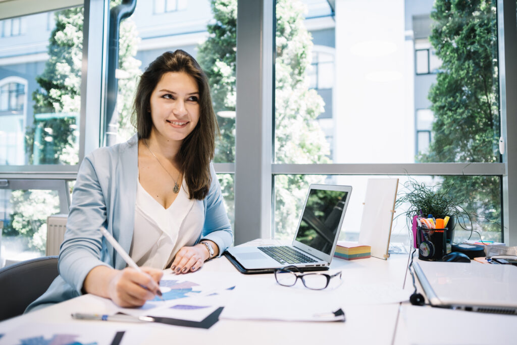 Smiling woman in her office sat at her desk, writing notes, with her laptop open and her glasses on her desk