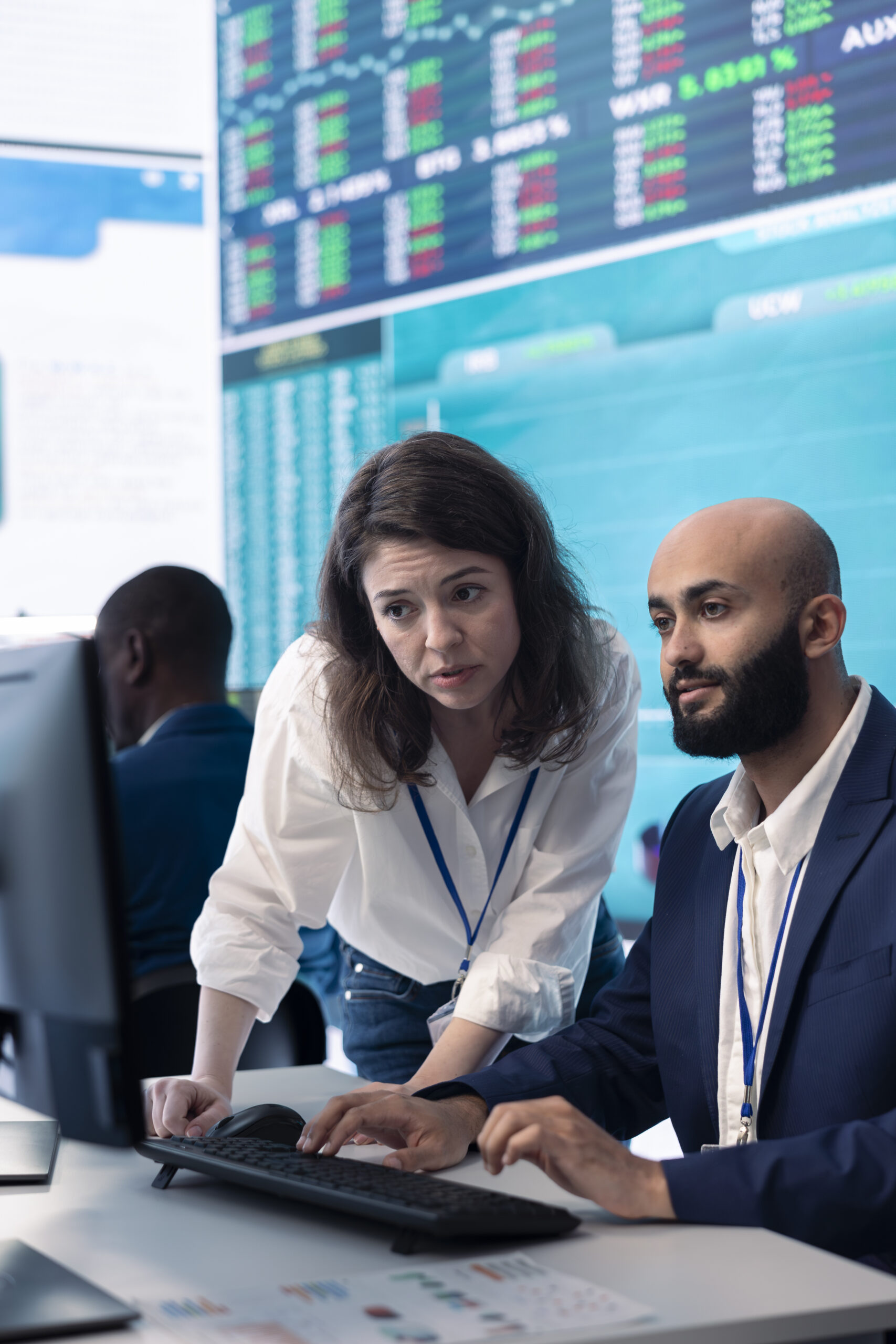 Manager helping out her team in the office at his desk with a huge live monitor behind them