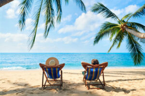 a couple sat on deck chairs on the beach with their arms folded behind their heads looking at the ocean under a palm tree