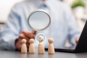 A man sat at a desk holding a magnifying glass over wooden figures with a tick symbol. Illustrating sucessful right to work checks.